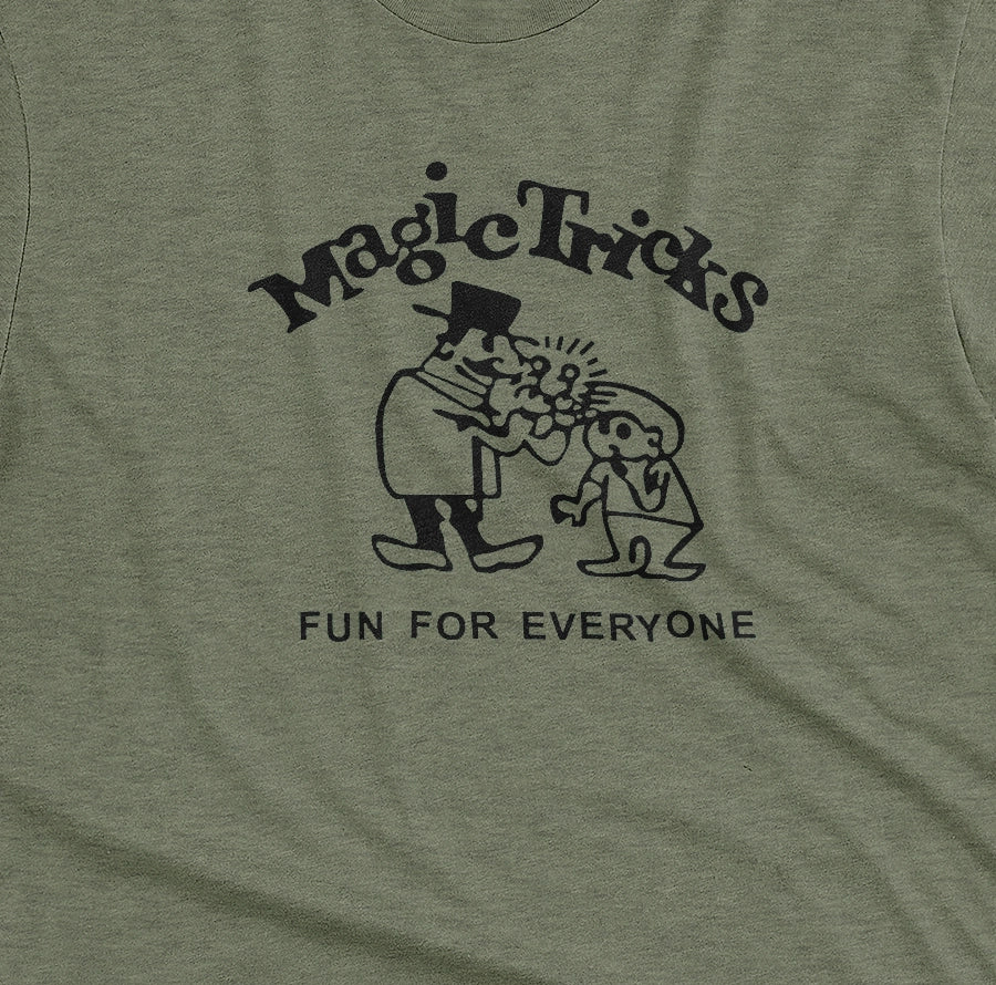 A green t-shirt with a black graphic print on the front that reads 'Magic Tricks' above a cartoon of a magician and a spectator, with 'FUN FOR EVERYONE' printed below.