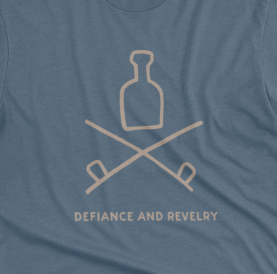 A blue t-shirt with a grey graphic of a bottle and crossed smoking pipes beneath it and the words defiance and revelry. The graphic is from an 1800s Australian convict tattoo.