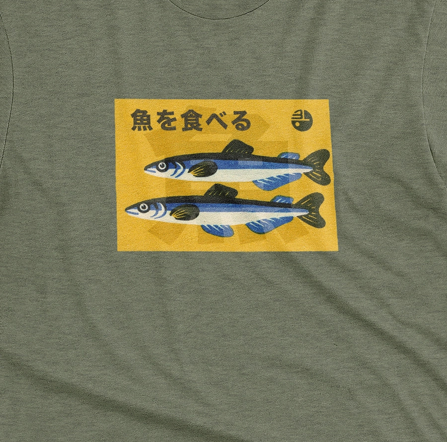 A green t-shirt with a rectangular graphic print on the chest showing two blue and silver fish on a yellow background with the Japanese text 'Eat Fish'.