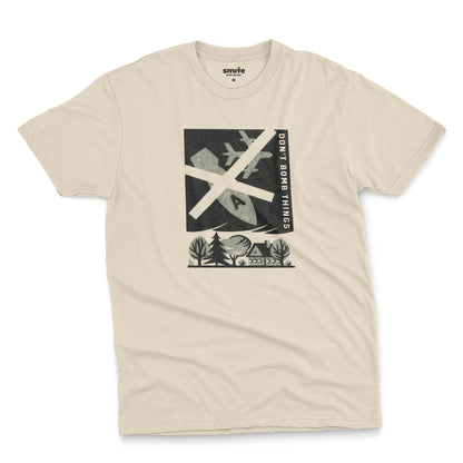 A beige t-shirt with a retro graphic print of an anti-war message featuring a cross, a plane, a house, and the text 'DON'T BOMB THINGS'.