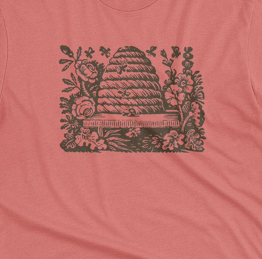 A coral pink t-shirt featuring a retro engraved graphic print of a beehive surrounded by flowers.