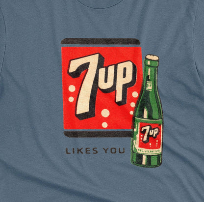 A blue t-shirt featuring a retro advertisement for 7up with the text '7up LIKES YOU'.
