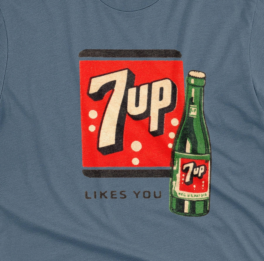 A blue t-shirt featuring a retro advertisement for 7up with the text '7up LIKES YOU'.