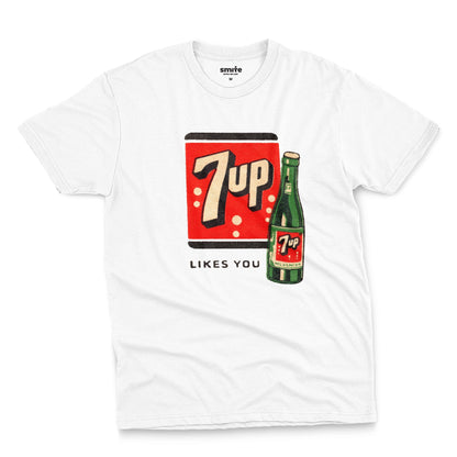 A white t-shirt featuring a retro advertisement for 7up with the text '7up LIKES YOU'.